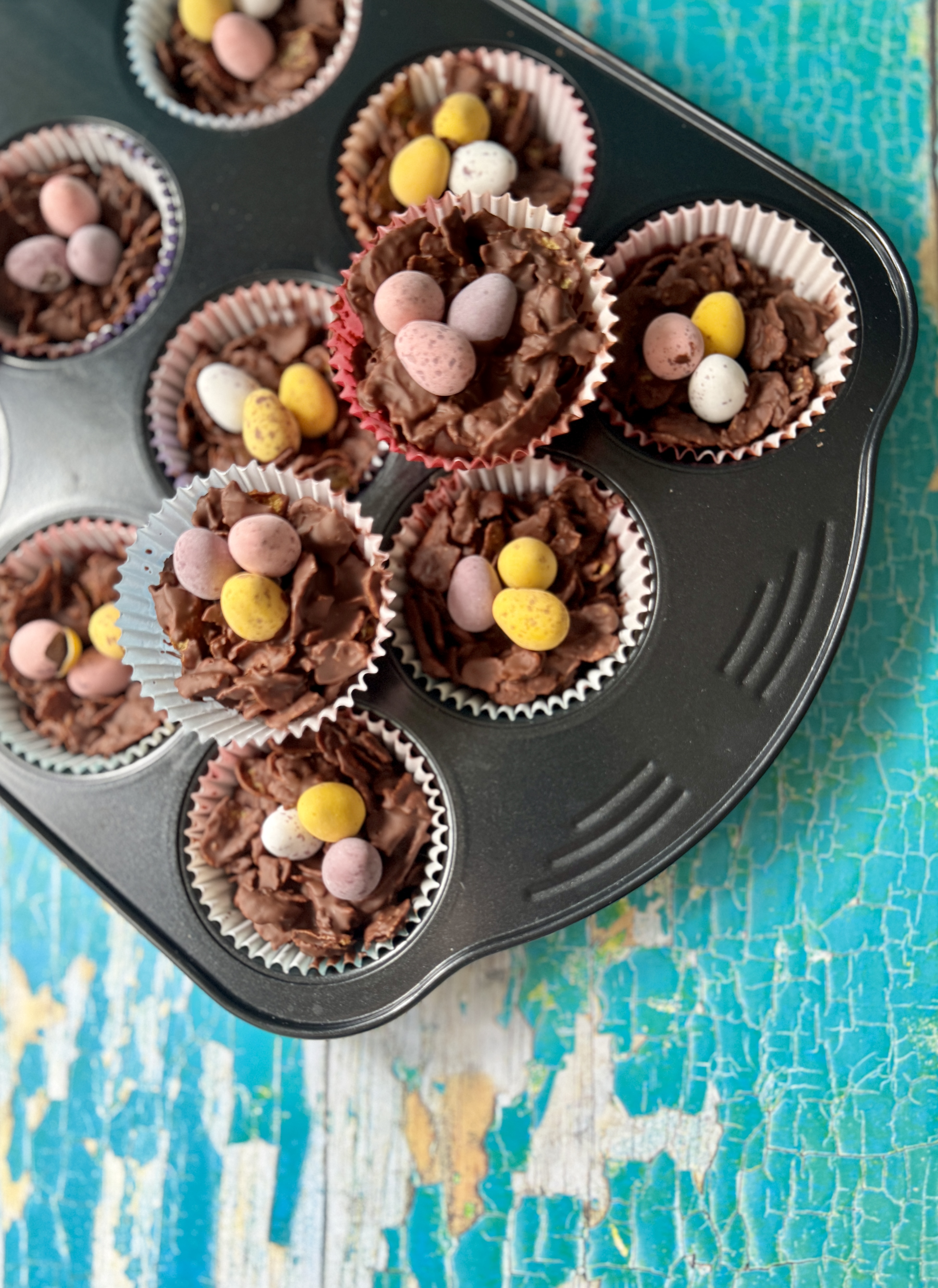Homemade Easter cornflake cakes in pastel paper cases, arranged in a cupcake tray on a rustic turquoise background with two cakes lifted for detail.